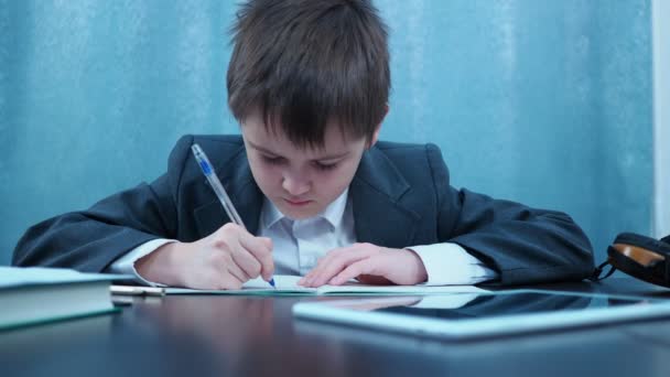 Un niño con una chaqueta escribe en un cuaderno y luego mira el marco y sonríe — Vídeo de stock