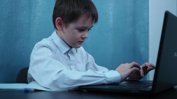 Een jongen in een shirt typt op de sleutels van een laptop die aan een tafel zit. draaien om te glimlachen naar de camera — Stockvideo