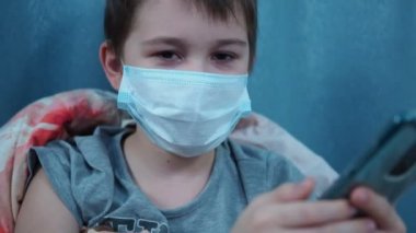 Tıbbi maskeli bir çocuk akıllı telefonuyla battaniyenin altında yatıyor.