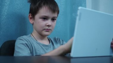 Tişörtlü bir çocuk masada otururken beyaz bir tabletle oynuyor.