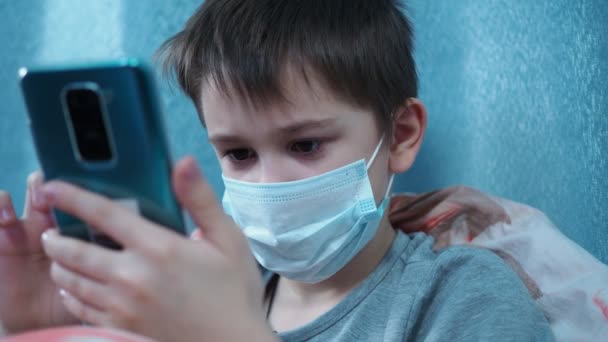 담요 밑에 누워 의료용 마스크를 쓰고 있는 한 소년이 스마트폰에 번호를 적는다 — 비디오
