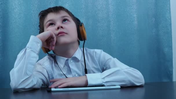 El muchacho en los auriculares escucha tranquilamente la lección sin mirar la tableta — Vídeos de Stock