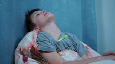 Yataktaki çocuk termometreyle ısıyı ölçer, döner ve kameraya gülümser.