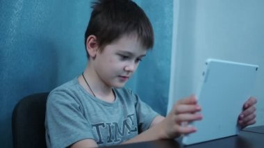 Tişörtlü bir çocuk sosyal ağlarda beyaz bir tablette masada oturuyor.