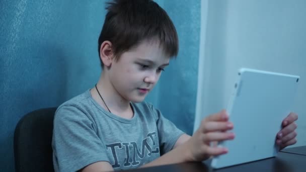 Ένα αγόρι σε ένα t-shirt γυρίζει μέσα από τα κοινωνικά δίκτυα σε ένα λευκό δισκίο κάθεται σε ένα τραπέζι — Αρχείο Βίντεο