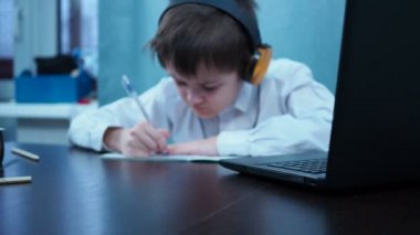 Gömlekli bir çocuk dizüstü bilgisayarın başına oturmuş deftere yazıyor. Çocuk odaklanamıyor.