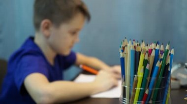 Çocuk masada oturan kalemlerle resim çizer. Oğlan odak dışı, kalemler ön planda.