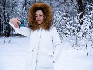 Karla kaplı ormanda beyaz ceketli bir kız telefonuyla fotoğraf çekiyor.
