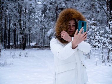 Karla kaplı ormanda beyaz ceketli bir kız telefonuyla fotoğraf çekiyor.