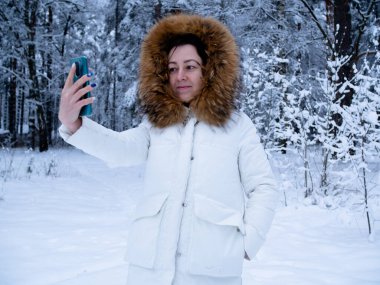 Karla kaplı ormanda beyaz ceketli bir kız telefonuyla fotoğraf çekiyor.