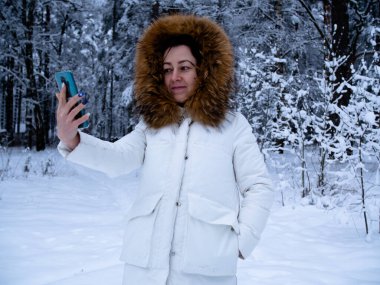 Karla kaplı ormanda beyaz ceketli bir kız telefonuyla fotoğraf çekiyor.