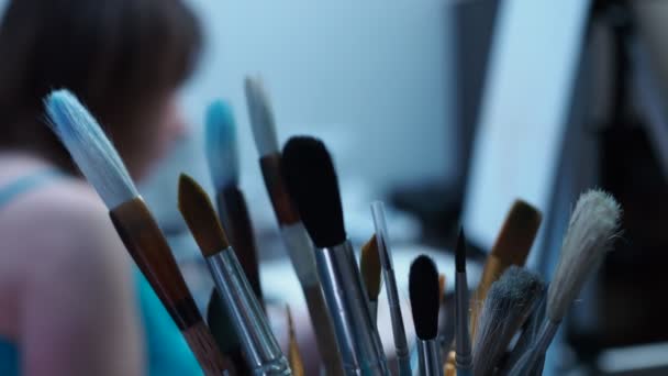 Un artista talentoso dibuja con un pincel sobre lienzo. vista desde detrás de las borlas. pinceles en foco, chica fuera de foco — Vídeos de Stock