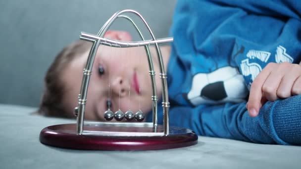 Um menino brinca com um pêndulo Newtons bolas deitado no divino — Vídeo de Stock