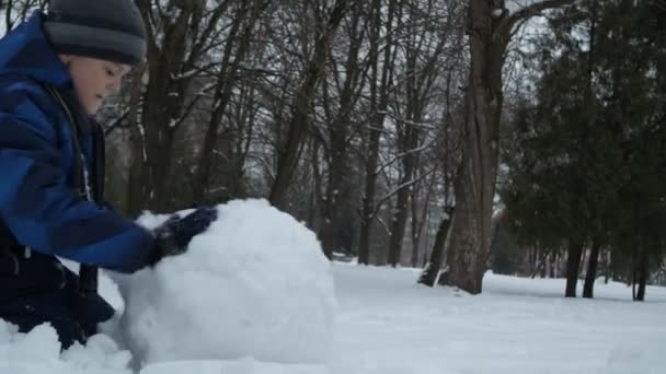 Karácsonyi órák. Egy kék kabátos fiú egy hógolyót gurít. — Stock videók