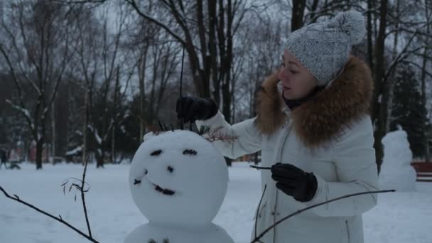 Karácsonyi órák. Egy fiatal nő hóembert díszít a parkban. — Stock videók