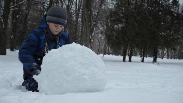 Karácsonyi órák. Egy kék kabátos fiú havat faragott a parkban. — Stock videók