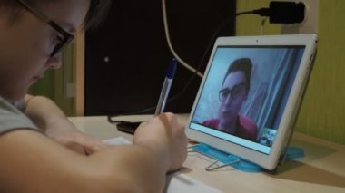 Ders online. Gözlüklü çocuk masada oturuyor, kameraya dönüyor ve gülümsüyor. Genç bir öğretmen programı açıklar.