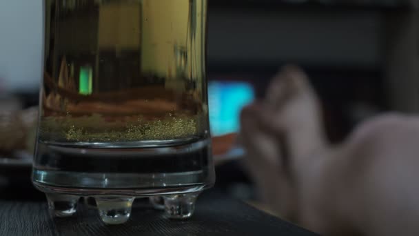 Beer bubbles in the glass in front of the TV. mens legs are out of focus — Wideo stockowe