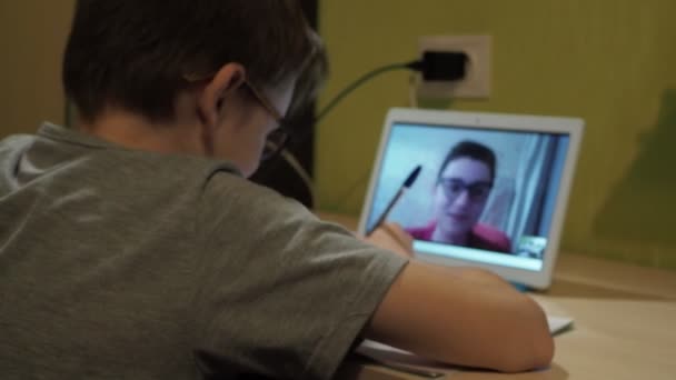 Aula online. um menino com óculos senta-se em uma mesa e escreve em um caderno. um jovem professor explica o programa — Vídeo de Stock