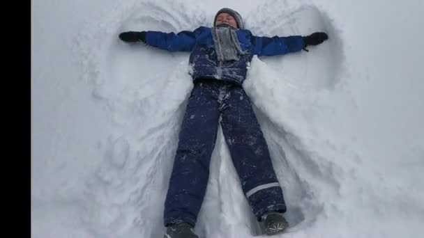 De jongen die op de sneeuw ligt beweegt zijn armen en benen. Een engel in de sneeuw portretteren. wintervermaak — Stockvideo