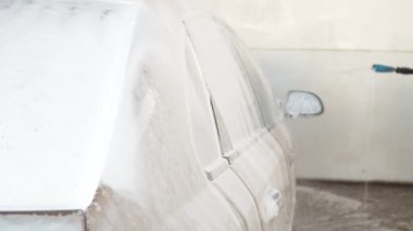 Araba, araba yıkamada köpükle doldurulur. Ceketli bir çocuk arabayı yıkar. Araba bej köpüklü.