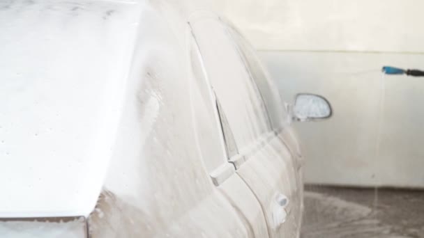 The car is poured with foam at the car wash. a boy in a jacket washes the car. the car is beige in foam — Stock Video