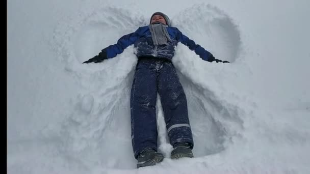 De jongen die op de sneeuw ligt beweegt zijn armen en benen. het beeld van een engel in de sneeuw. wintervermaak — Stockvideo