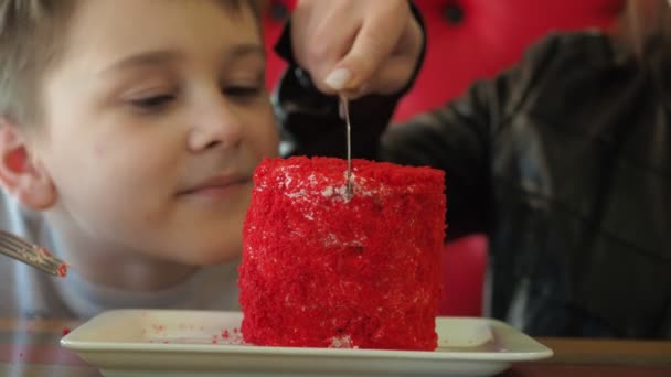Chłopiec jest pokrojony w tort na talerzu. Czerwony gąbczasty tort w centrum uwagi, chłopcze nieostry. Emocje zaskoczenia, szczęście na twarzy dziecka — Wideo stockowe
