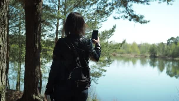 Una mujer joven con una chaqueta de cuero se encuentra en la orilla del lago y toma fotos en su teléfono inteligente. viaje — Vídeos de Stock