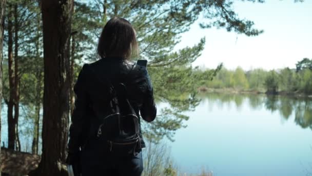 Uma jovem mulher em uma jaqueta de couro fica na margem do lago e tira fotos em seu smartphone — Vídeo de Stock