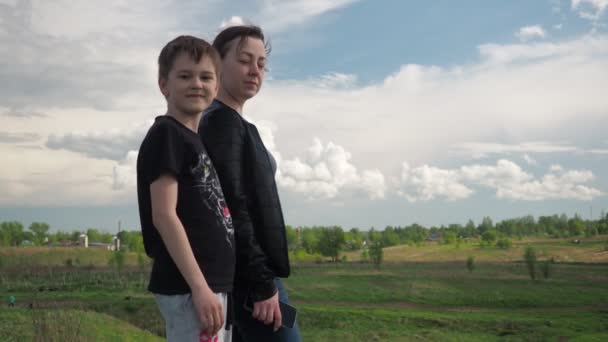 Um rapaz com uma jovem mãe numa colina a olhar para a câmara. família feliz. o menino acena sua mão e salta — Vídeo de Stock