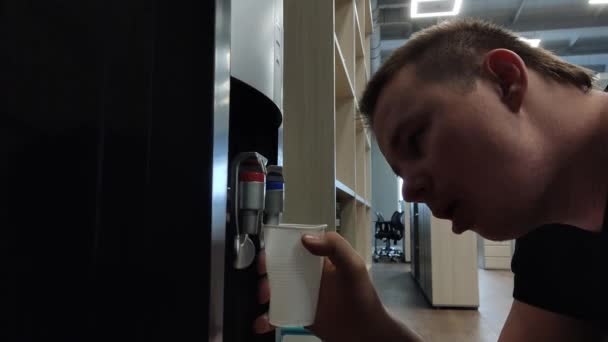 En man med baksmälla häller vatten från en kylare på ett stort kontor. hård arbetsdag — Stockvideo