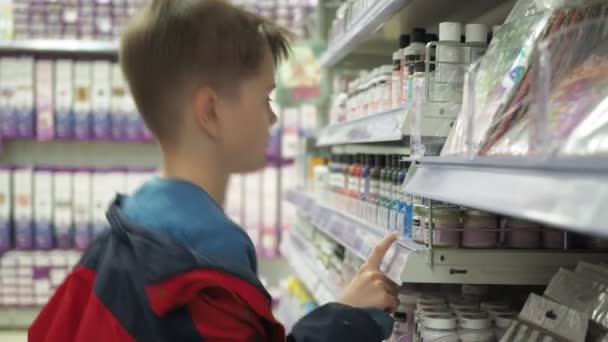 Un chico con una chaqueta roja elige pintura en el mostrador. miramos los frascos con líquidos — Vídeo de stock