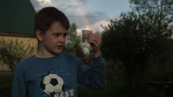 Ένα αγόρι συλλέγει σταγόνες βρόχινου νερού σε ένα ποτήρι ενώ στέκεται πάνω σε ένα φόντο ουράνιου τόξου σε ένα οικόπεδο της χώρας — Αρχείο Βίντεο