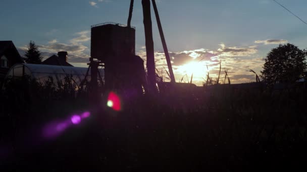 Un hombre con chaqueta está caminando en una parcela de granja. el sol golpea la cámara con un rayo formando purpurina. el hombre lleva puesta una camiseta con gorra — Vídeo de stock