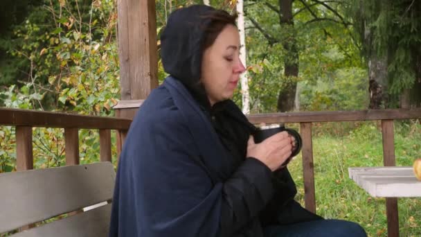 Una joven con una chaqueta negra está sentada bebiendo café caliente en el parque en la mesa del cenador. otoño frío. Está resfriada. secreción nasal. calentando con una bebida caliente — Vídeo de stock