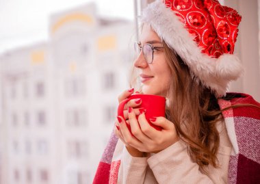 Noel Baba şapkalı güzel genç kız elinde kırmızı bir fincan çayla pencereden dışarı bakar, Noel ve yeni yıl temasını hayal eder.