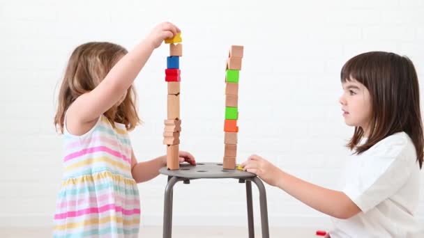 Сестра 8 лет и девочка 4 лет строят башню из ярких деревянных кубиков, играют, улыбаются, сидят дома на полу на белом фоне — стоковое видео