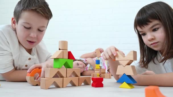 Adolescente menino 11 anos e sua irmã 8 anos e menina 4 anos crianças construir uma torre de cubos de madeira brilhante jogar construtor, sorrir, sentar no chão em um fundo branco em casa — Vídeo de Stock