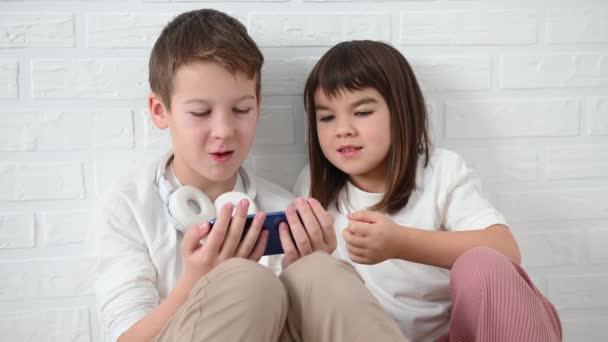 Adolescente, menino 11 anos com sua irmã 8 anos emocionalmente jogar um telefone azul, ficar com raiva, sorrir, sentar no chão em um fundo branco — Vídeo de Stock