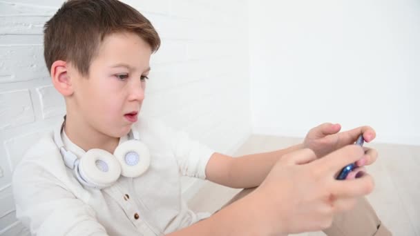 Tiener, jongen 11 jaar emotioneel spelen een blauwe telefoon, boos, glimlach, zitten op de vloer op een witte achtergrond — Stockvideo