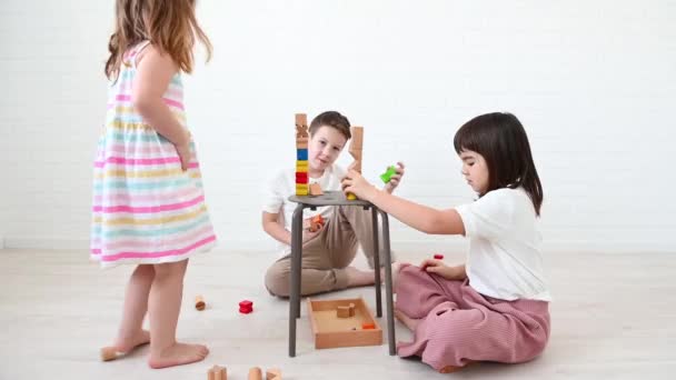 Adolescente menino 11 anos e sua irmã 8 anos e menina 4 anos crianças construir uma torre de cubos de madeira brilhante jogar construtor, sorrir, sentar no chão em um fundo branco em casa — Vídeo de Stock