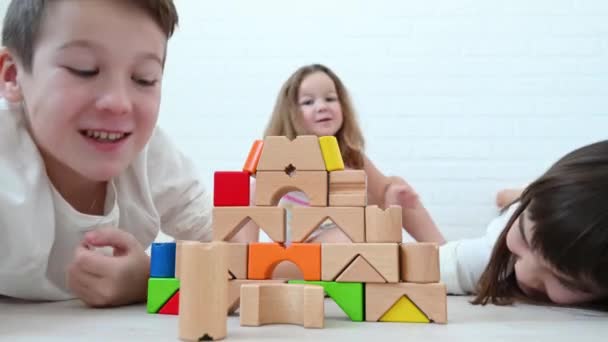 Nastolatek chłopiec 11 lat i jego siostra 8 lat i dziewczyna 4 lat dzieci zbudować wieżę z jasnych drewnianych kostek grać konstruktor, uśmiech, siedzieć na podłodze na białym tle w domu — Wideo stockowe
