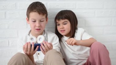 Genç, 11 yaşında kız kardeşiyle birlikte. Duygusal olarak 8 yaşında. Mavi bir telefon çalıyor, kızıyor, gülümsüyor, beyaz bir arka planda yerde oturuyor.