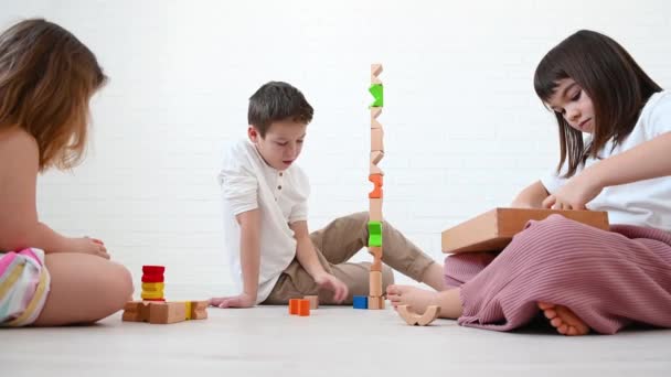 3人の子供ティーンエイジャーの男の子11歳と彼の妹8歳と女の子4歳の子供たちは 明るい木製のキューブプレイコンストラクター 笑顔から塔を構築し 白い背景に床に座って — ストック動画