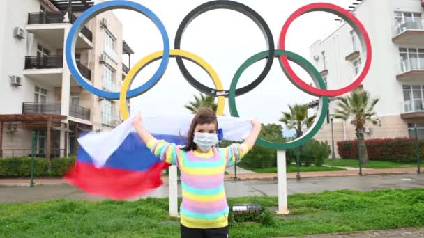 06,04,2021 Rusia, Sochi o persoană fetiță amuzantă într-o mască medicală stă cu steagul Rusiei lângă inelele olimpice tema jocurilor olimpice — Videoclip de stoc
