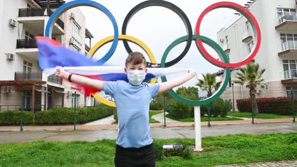 06,04,2021 Oroszország, Szocsi egy ember kis vicces fiú orvosi maszkban áll az oroszok zászlajával az olimpiai gyűrűk mellett a téma az olimpiai játékok — Stock videók