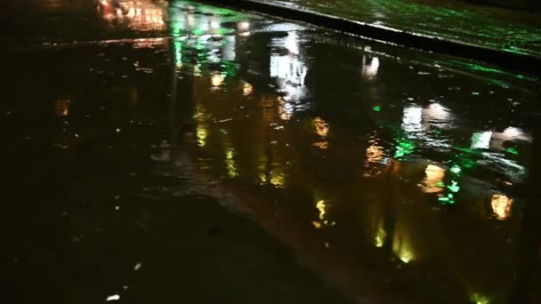 Ночной дождь в городе на дороге в лужах отражаются разноцветные огни — стоковое видео
