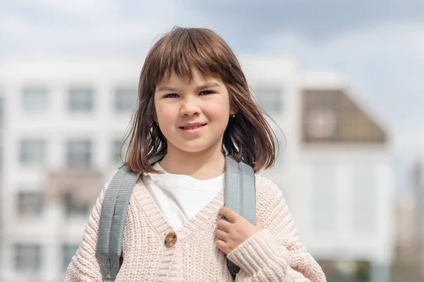 Chica feliz una colegiala de 8 años de apariencia europea con una mochila está caminando en el patio de la escuela por la tarde en la calle mirando la cámara de cerca —  Fotos de Stock
