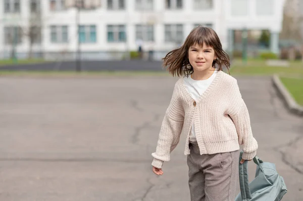 Ευτυχισμένο κορίτσι, μια μαθήτρια με 8 χρόνια ευρωπαϊκής εμφάνισης με ένα σακίδιο περπατά στην αυλή του σχολείου το απόγευμα στο δρόμο κοιτάζοντας από κοντά την κάμερα — Φωτογραφία Αρχείου